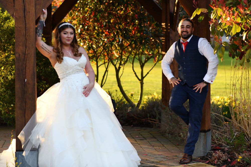Bride in flowing white gown poses under wooden pergola with autumn foliage backdrop.