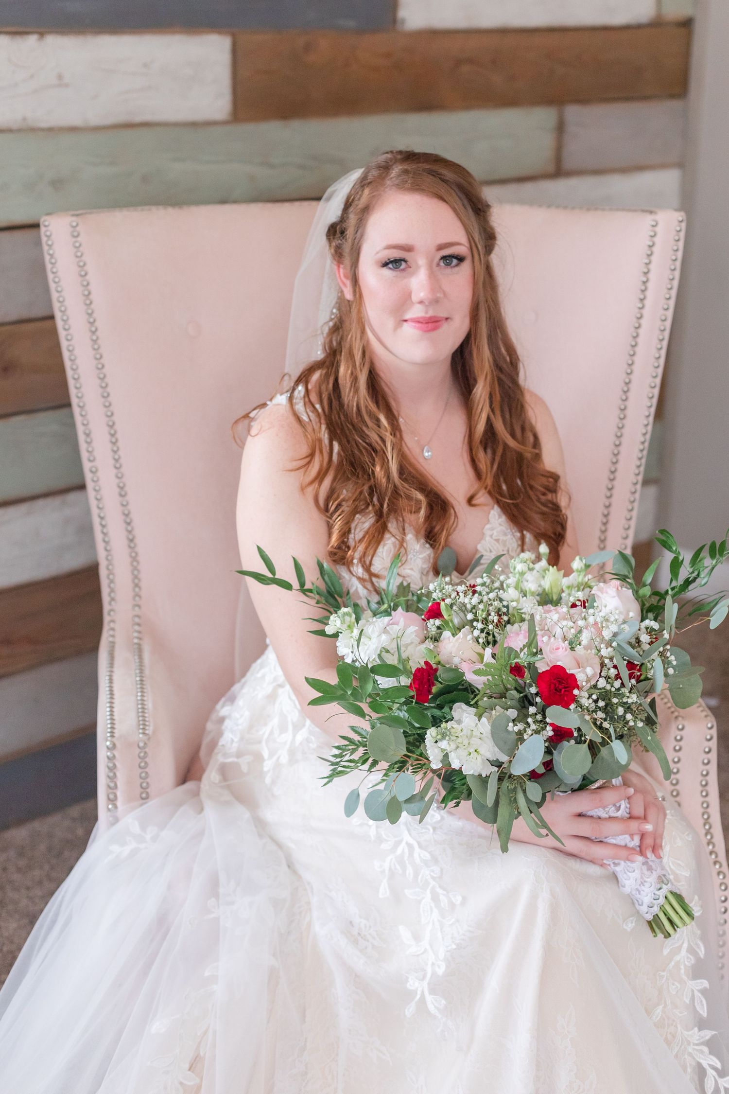A white wedding bouquet with red roses rests on a bride's lap in an upholstered chair.