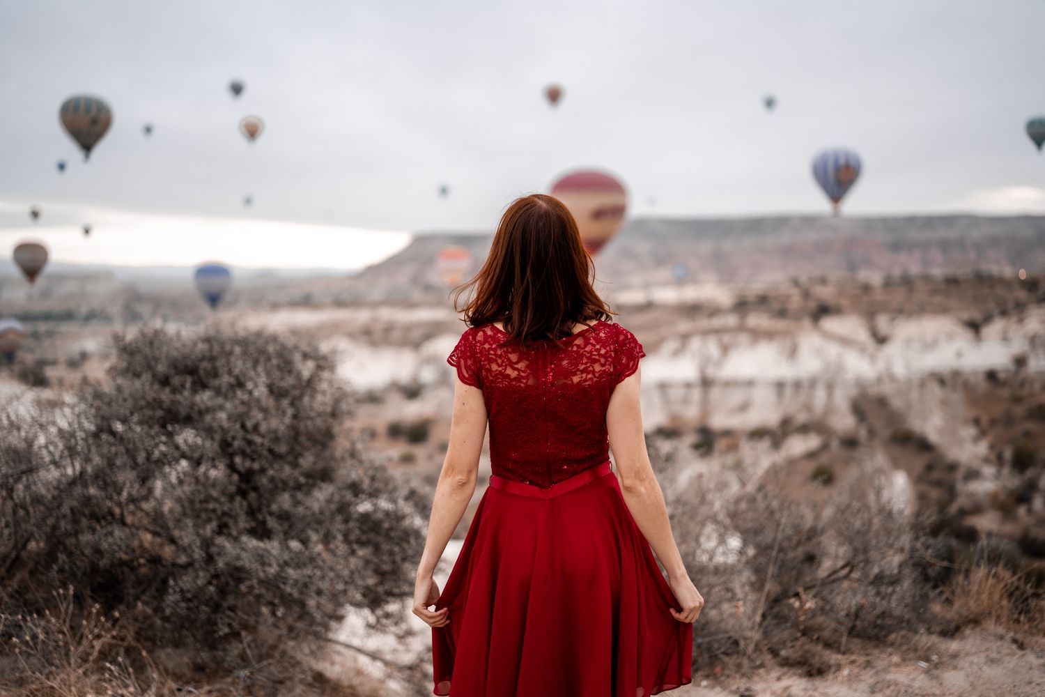 Woman in red dress views hot air balloons floating over desert landscape.