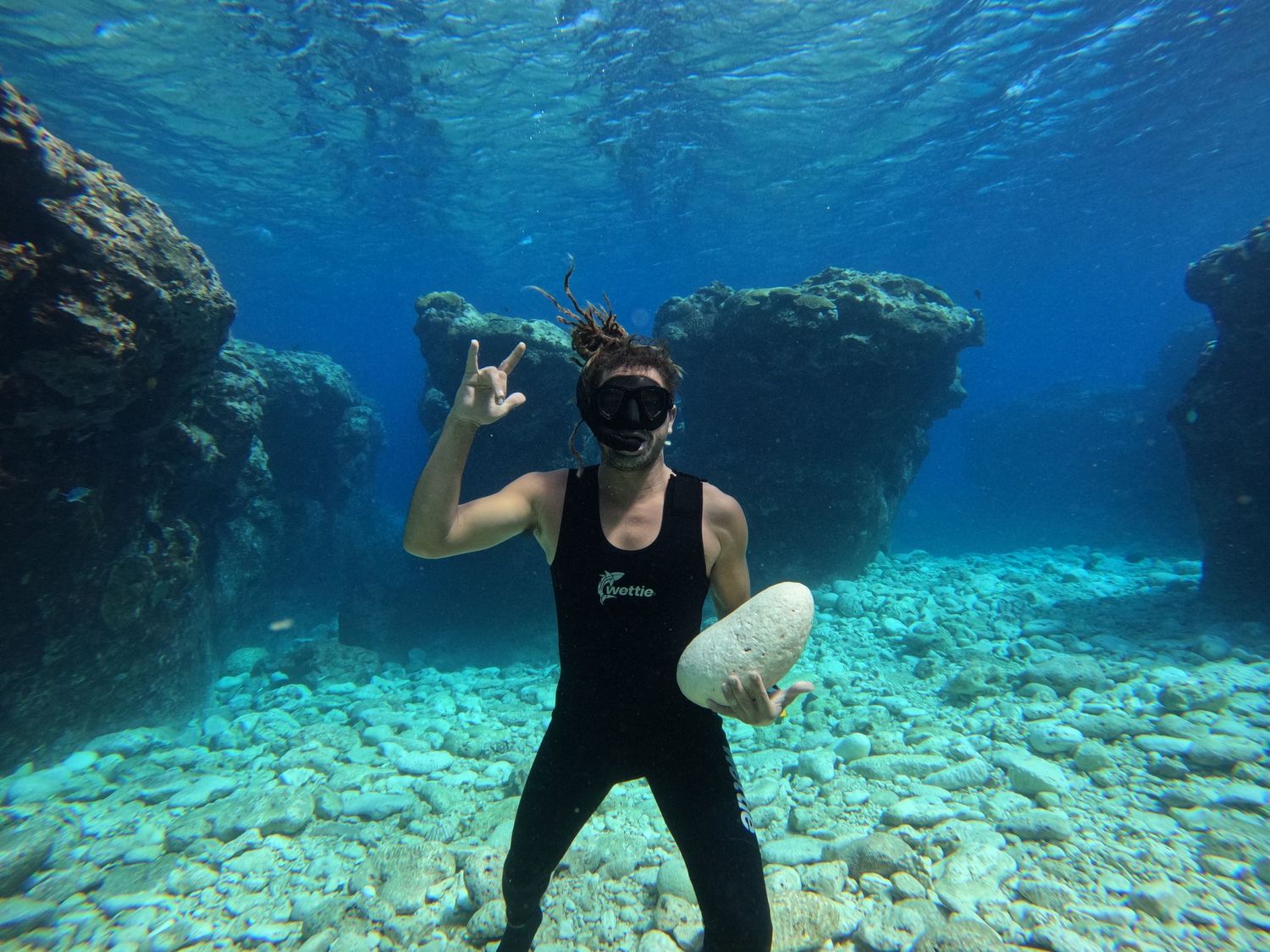 Diver with snorkel gear poses underwater near coral reef formations in clear blue water.