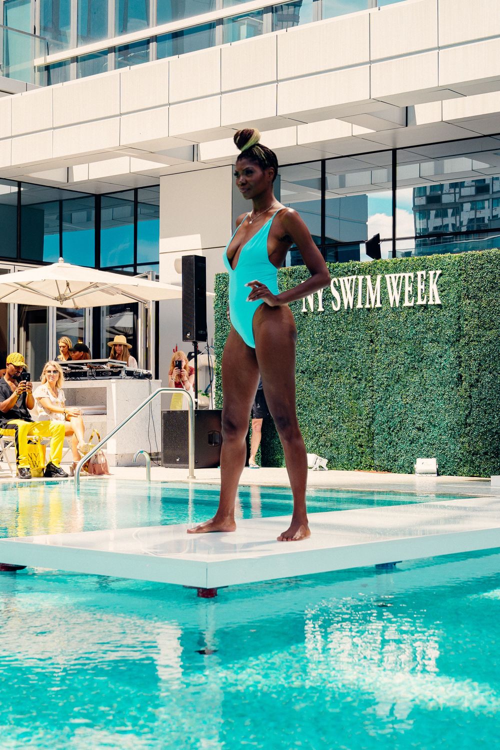 A summer pool scene with someone diving into bright turquoise water surrounded by glass railings and greenery.