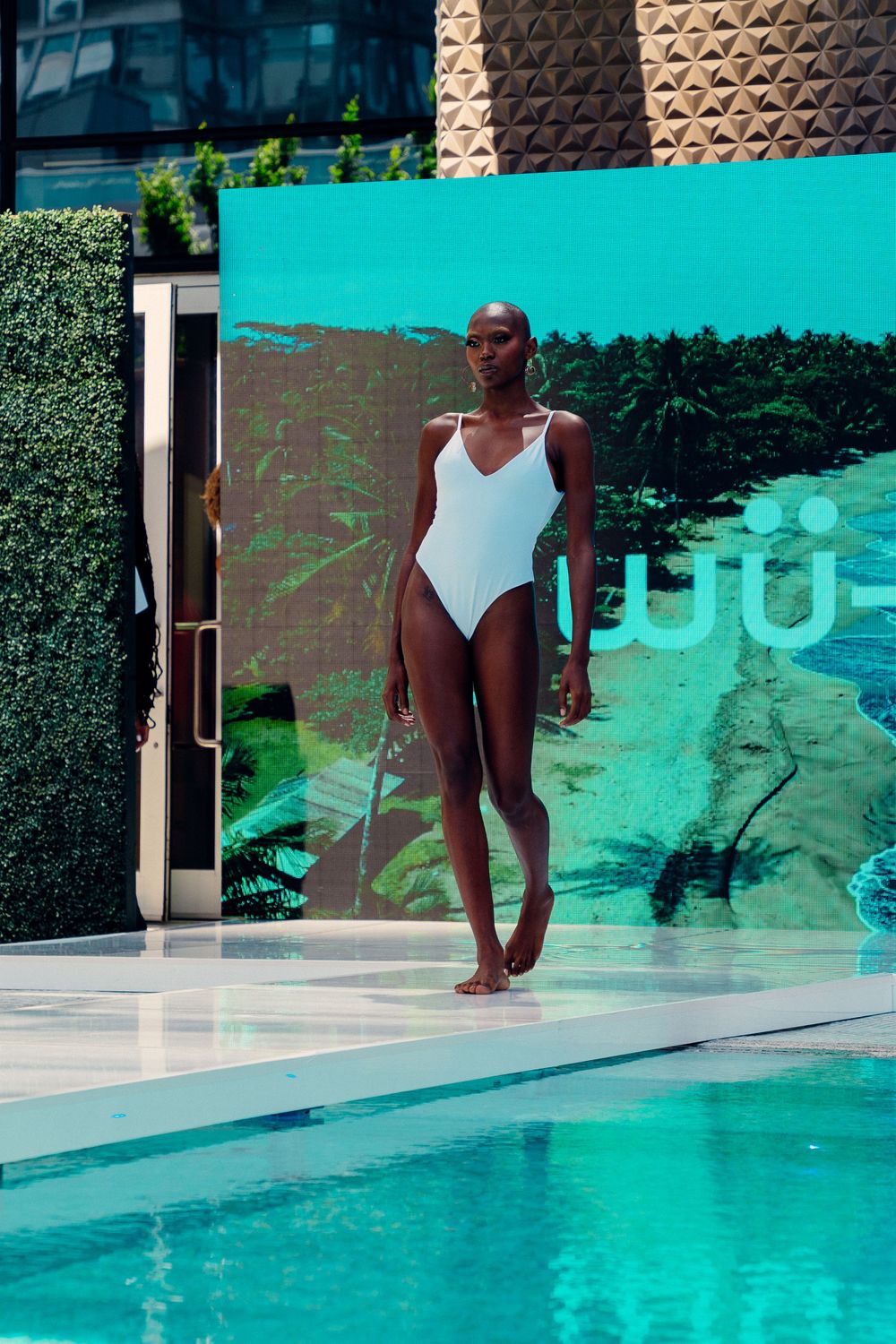 Athletic figure in a white swimsuit stands by a turquoise pool on a sunny day with greenery in the background.