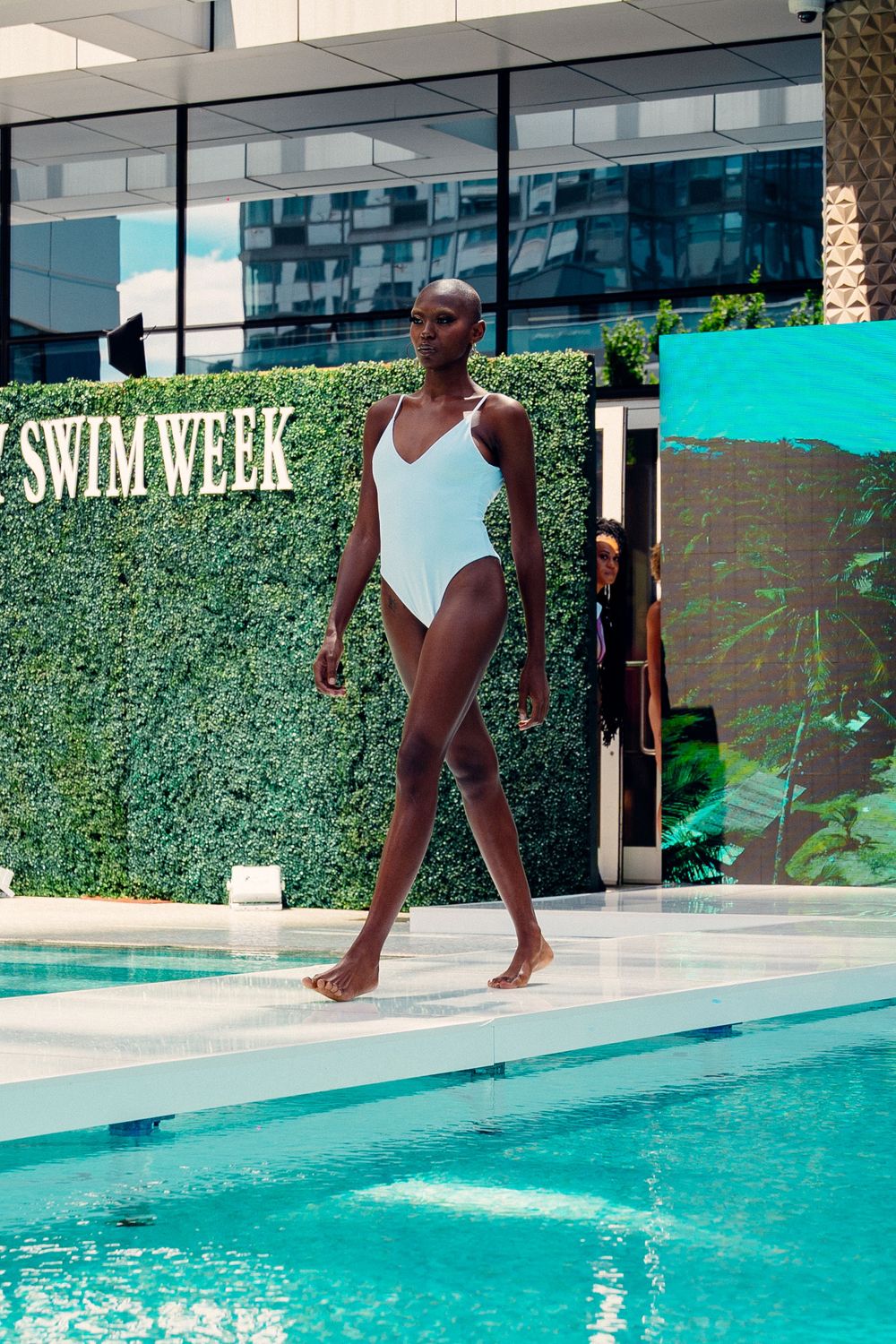 A person in a white swimsuit approaching the edge of a swimming pool with a green hedge backdrop.