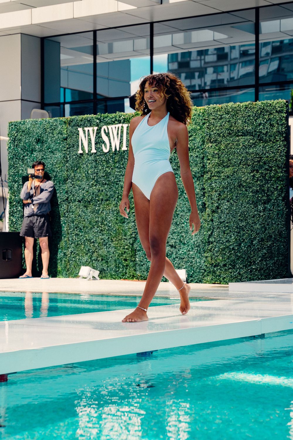 A swimming pool scene with turquoise water surrounded by modern architecture and landscaped hedges.