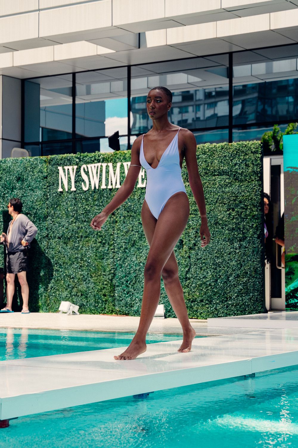 A person in a white swimsuit walking along the edge of a modern swimming pool with hedge walls.