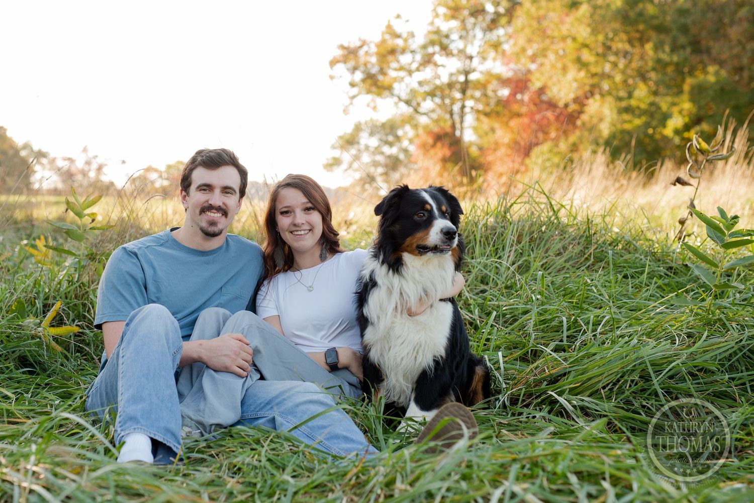 A couple sits with their Australian Shepherd dog in a sunny field during golden hour.