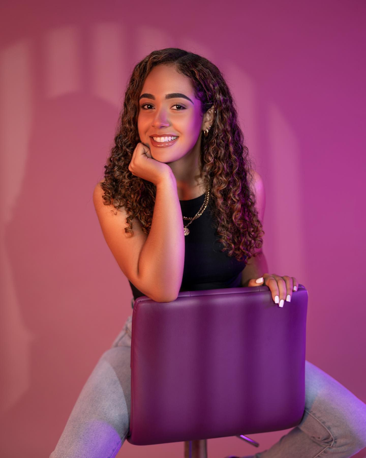 Portrait with purple chair against pink backdrop showing joyful expression.