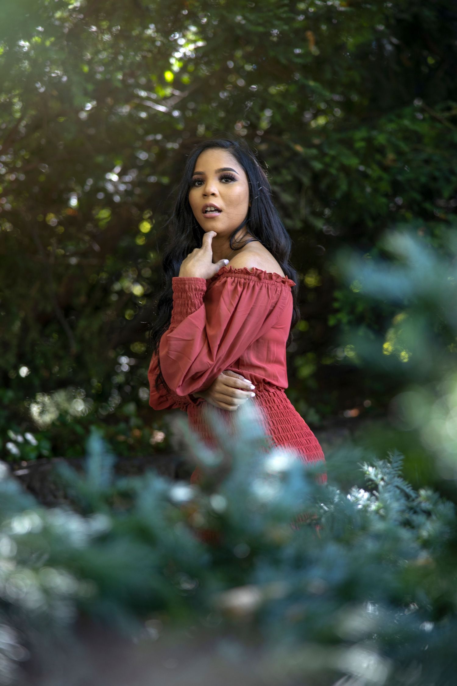 Portrait in red off-shoulder top surrounded by greenery with soft natural lighting.