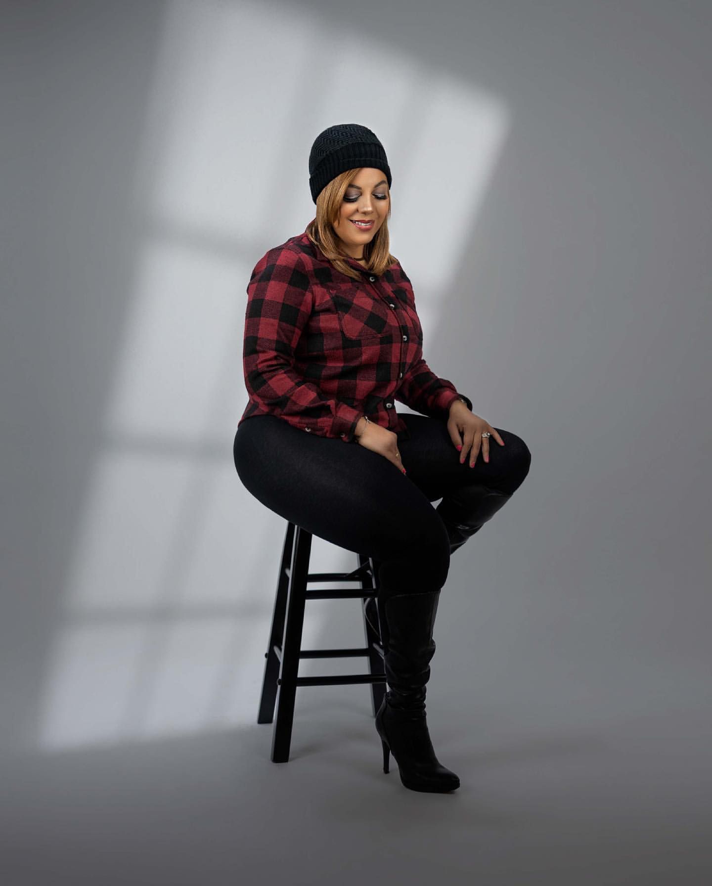 Person in plaid shirt and beanie poses on stool against grey studio backdrop with dramatic lighting.