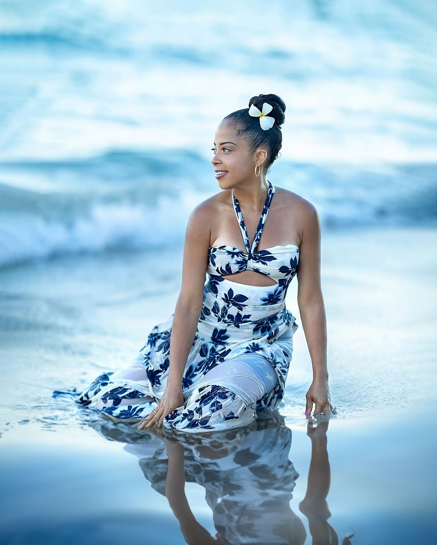Woman in floral print dress sits in shallow ocean water at sunset.