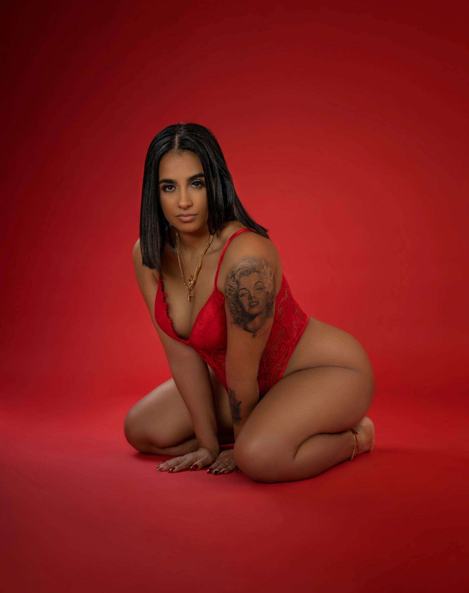 Artistic boudoir photo with red backdrop featuring model in red lingerie with visible tattoo.