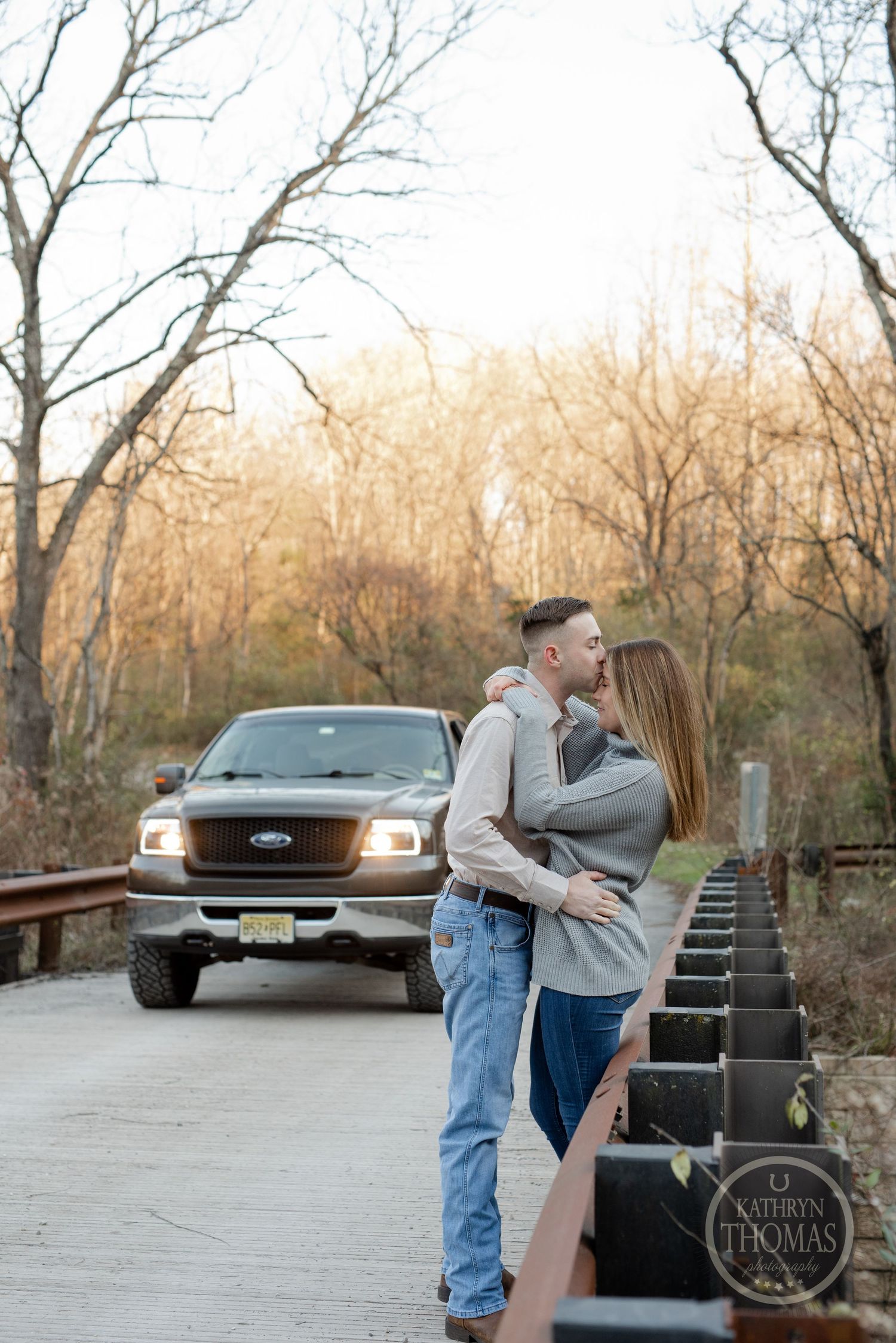 Couple embracing on a wooden bridge with a pickup truck parked in the autumn landscape.