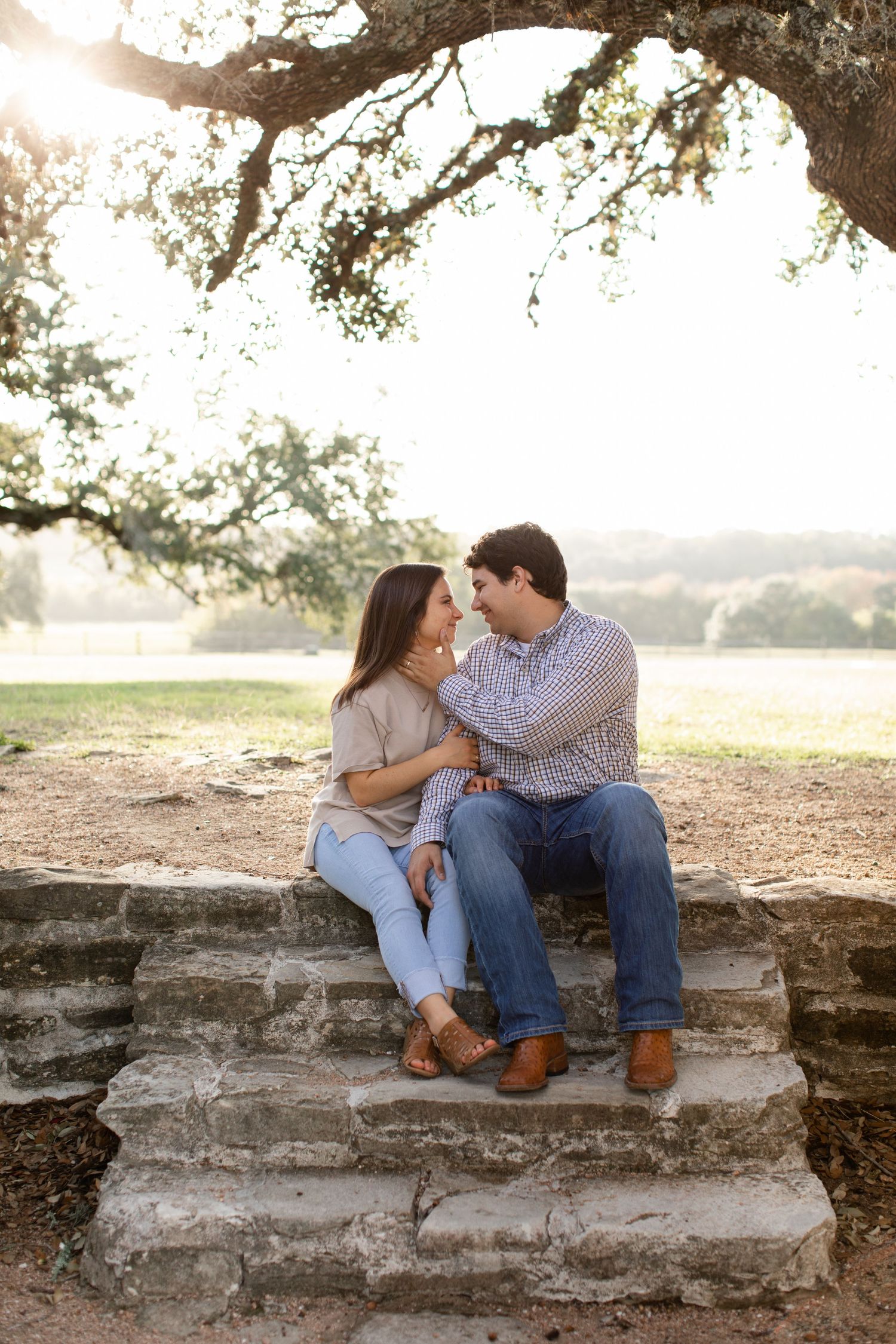 Romantic couple sitting close on stone steps under a tree branch at sunset in the countryside.
