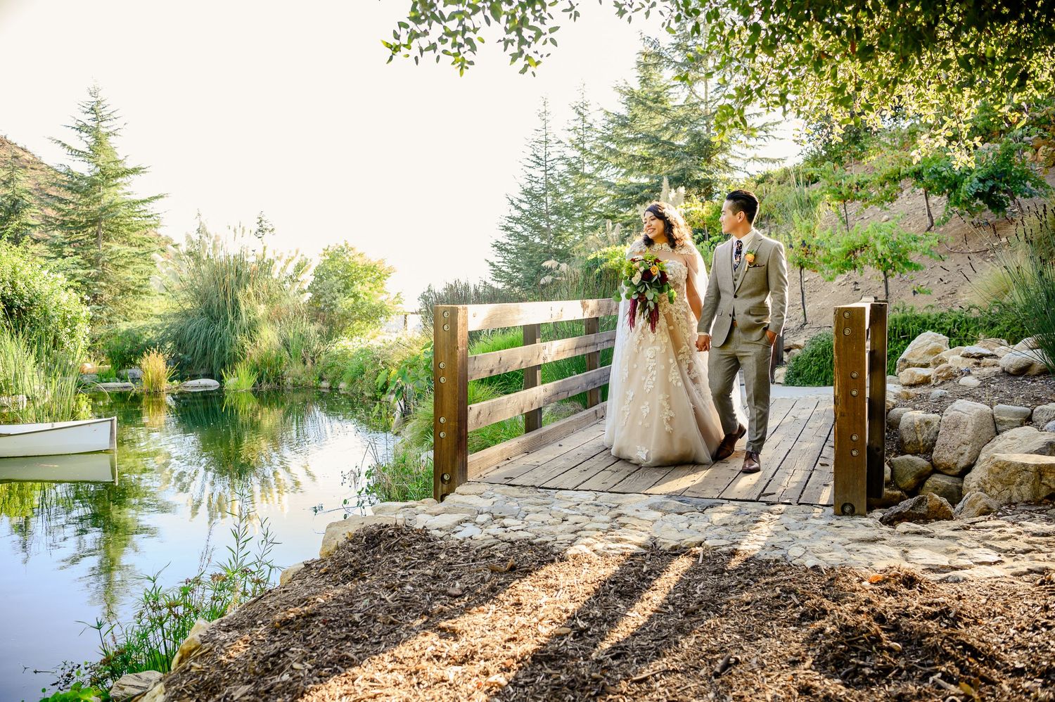 Couple standing on wooden footbridge over scenic pond with trees and mountains in background.