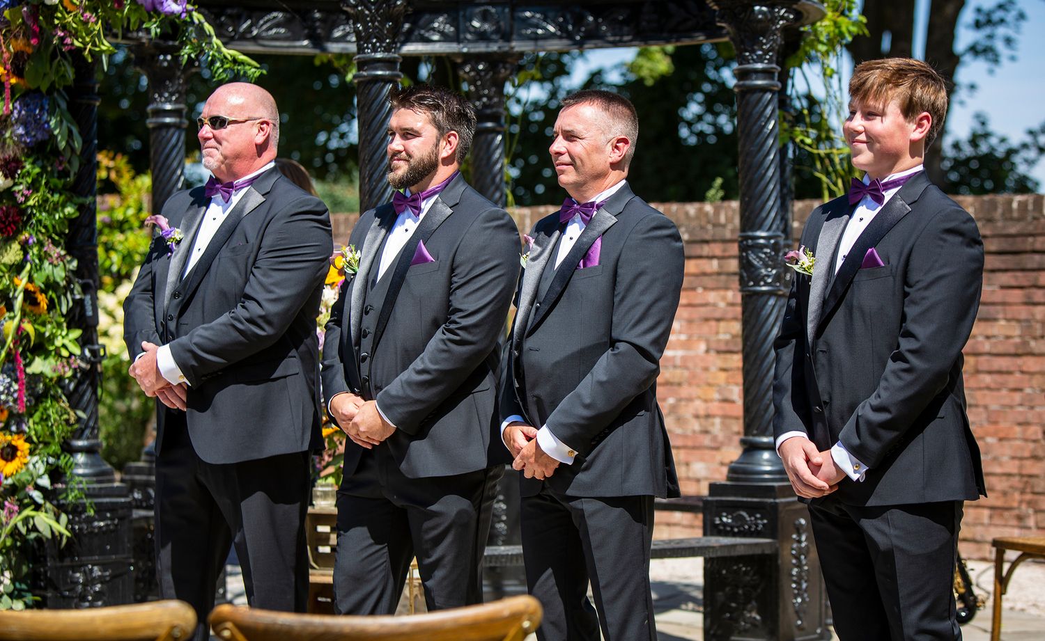 Groomsmen in black suits standing in line outdoors during a wedding ceremony.