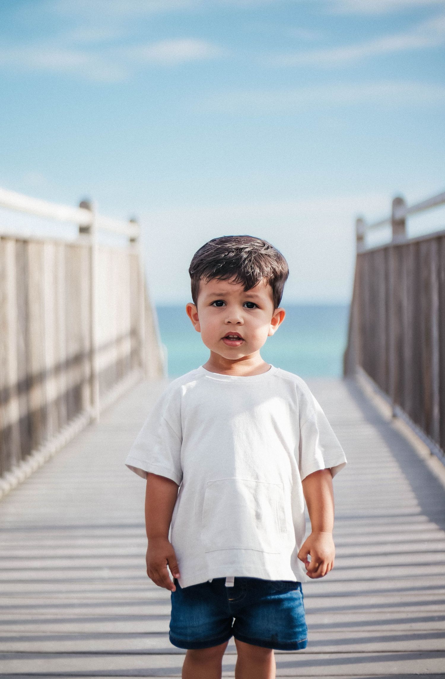 Small child in white t-shirt and blue shorts stands on wooden boardwalk by ocean.