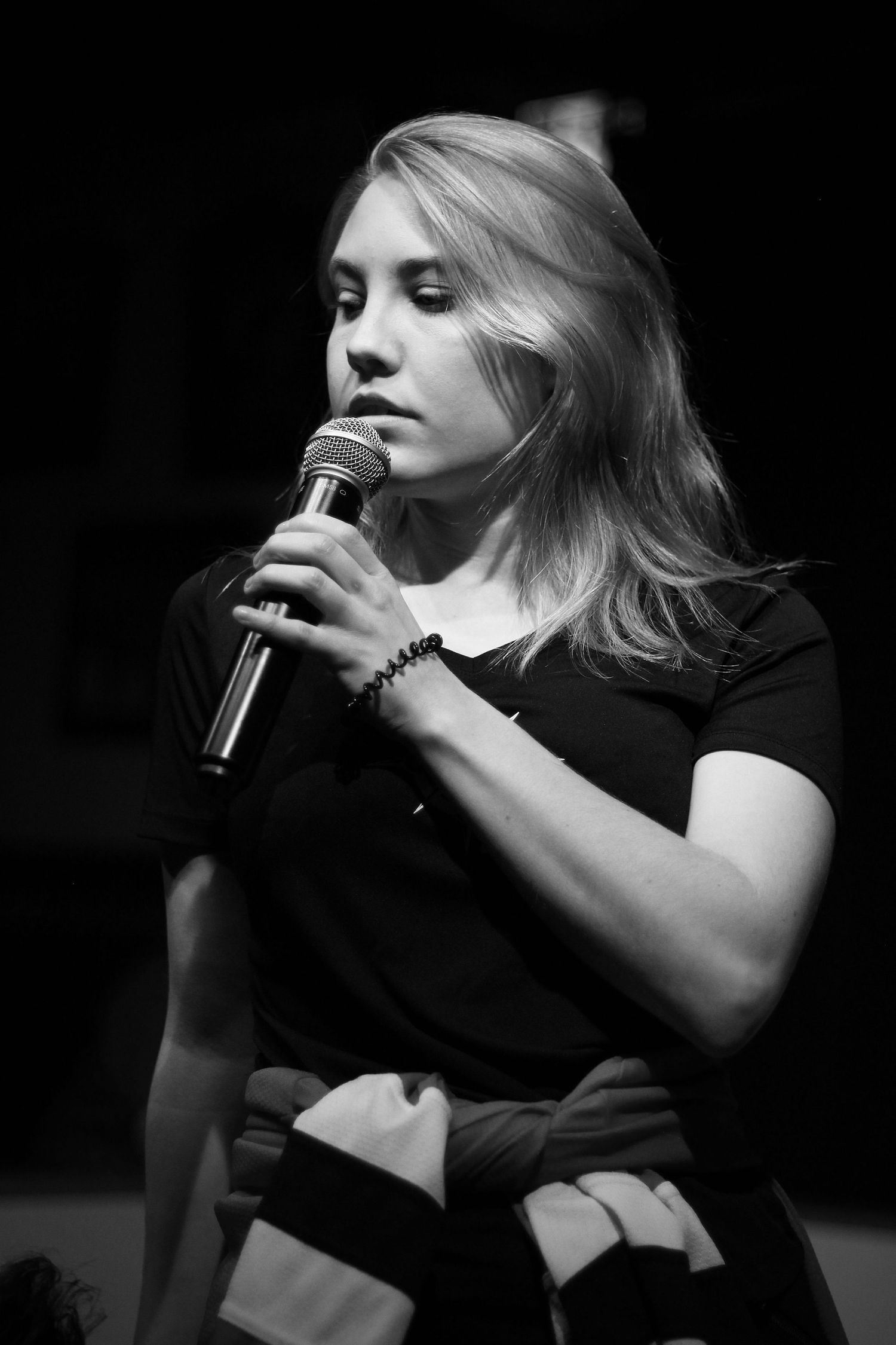 Young blonde woman holding a microphone, looking down thoughtfully in black and white photography.