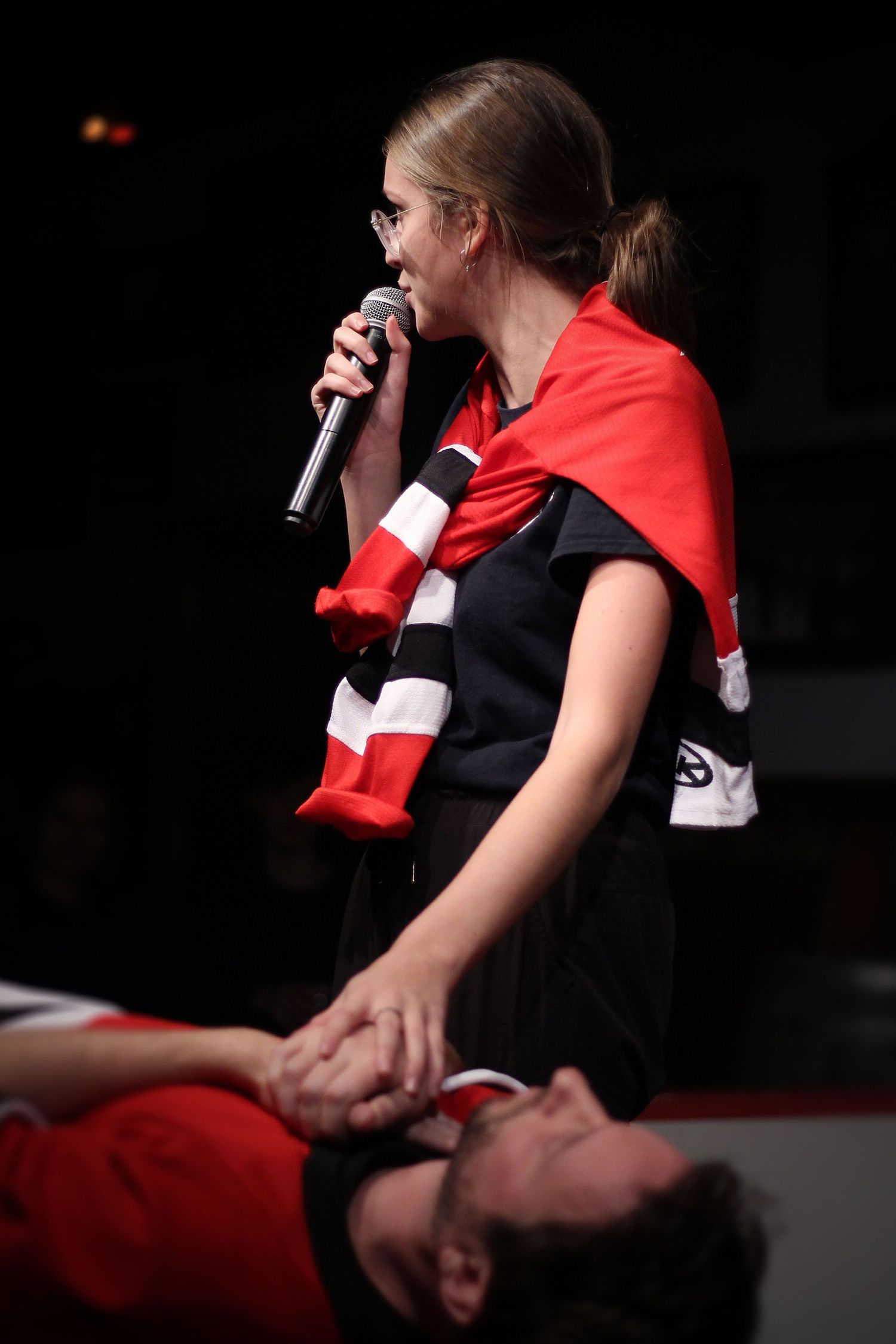 Woman wearing red scarf singing into microphone while standing over person lying on stage.