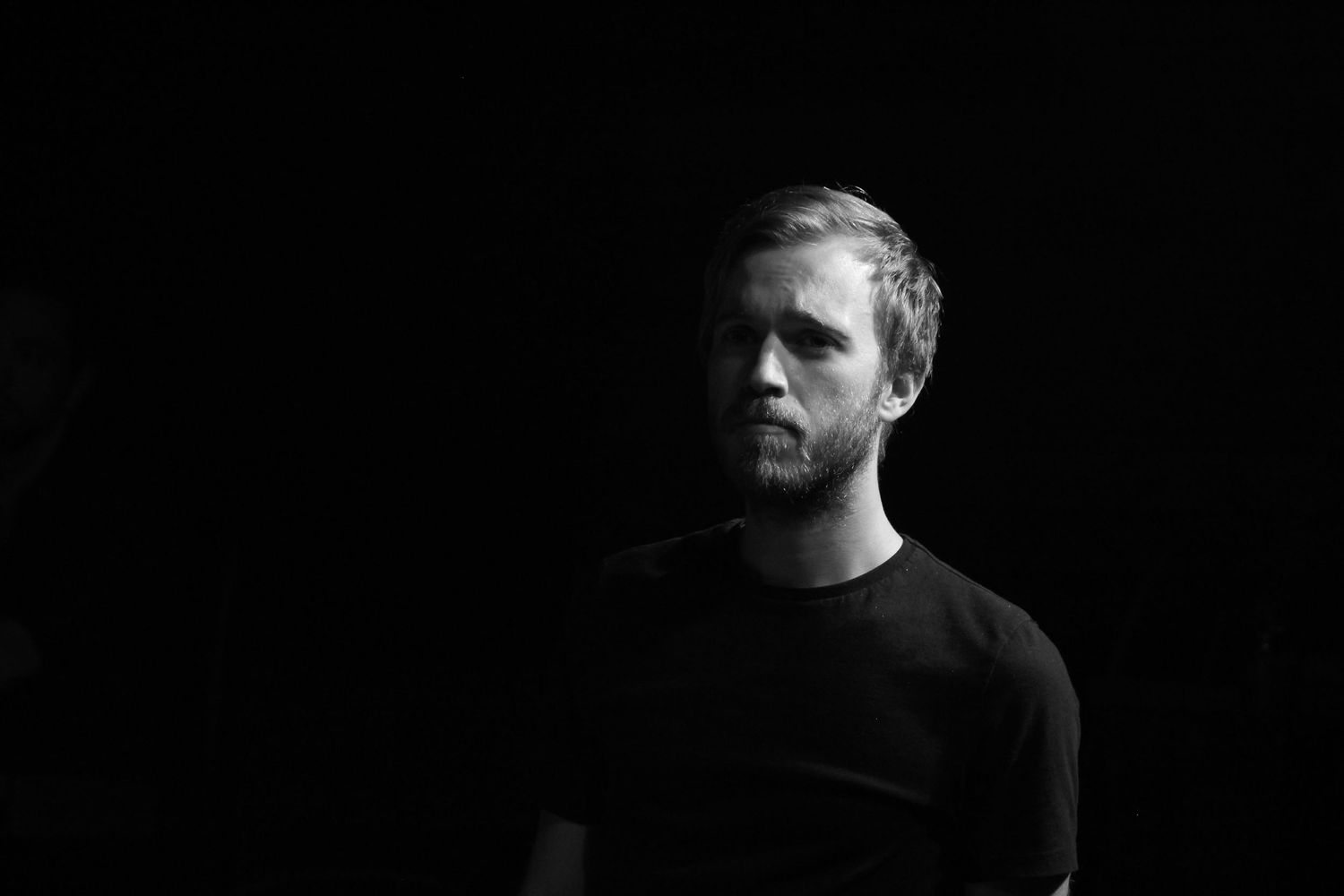 Black and white portrait of a bearded man in a dark sweater against a dark background.