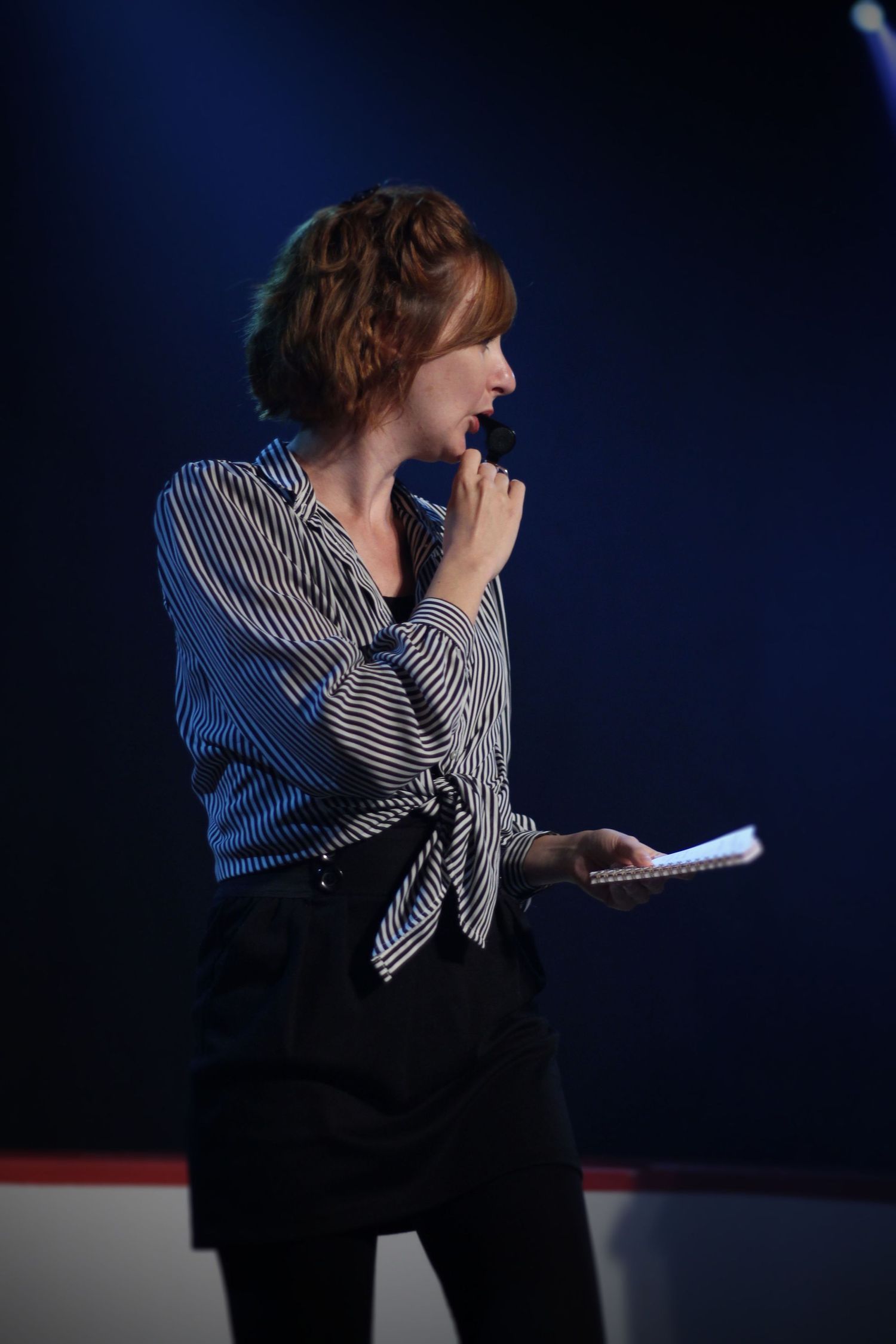 Woman in striped blouse holding microphone and notepad on a blue-lit stage during an event.