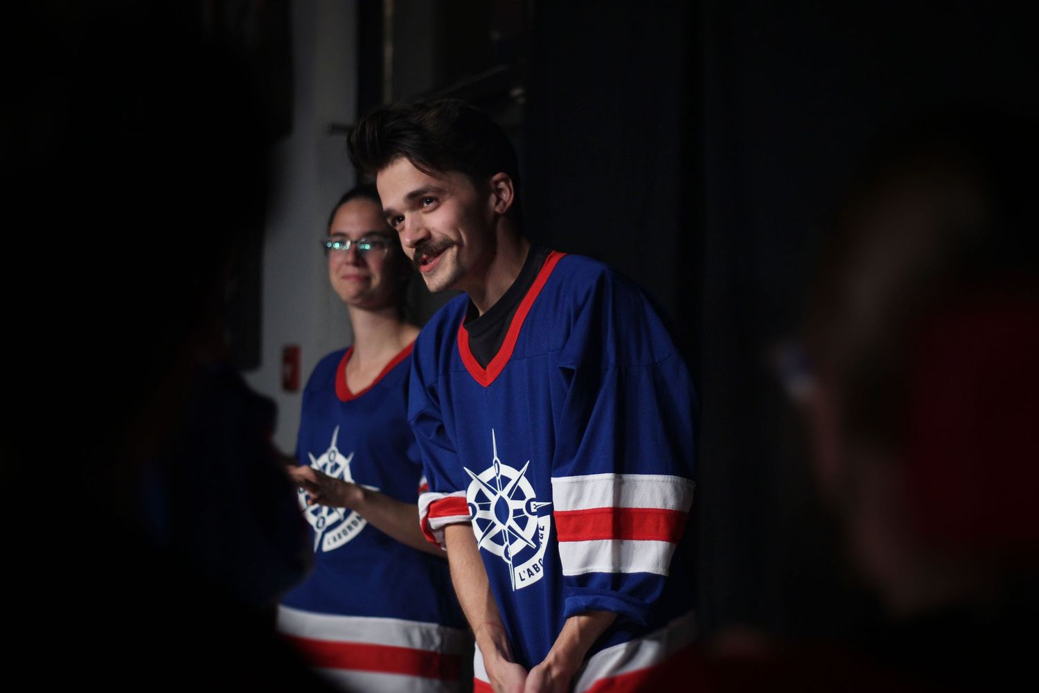 Two people wearing blue hockey jerseys smiling in a dimly lit venue.