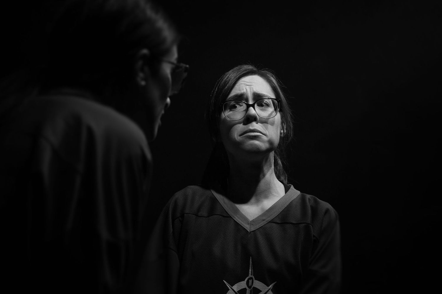 Black and white photo of a woman wearing glasses looking up at another person in dim lighting.