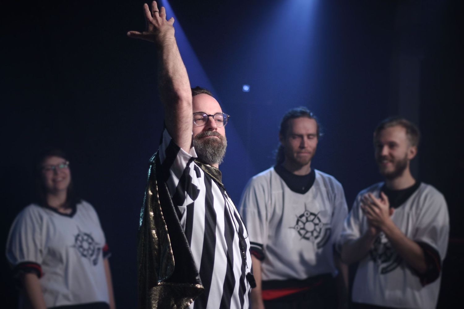 Man in black and white striped jacket raising hand on stage with teammates applauding behind him.