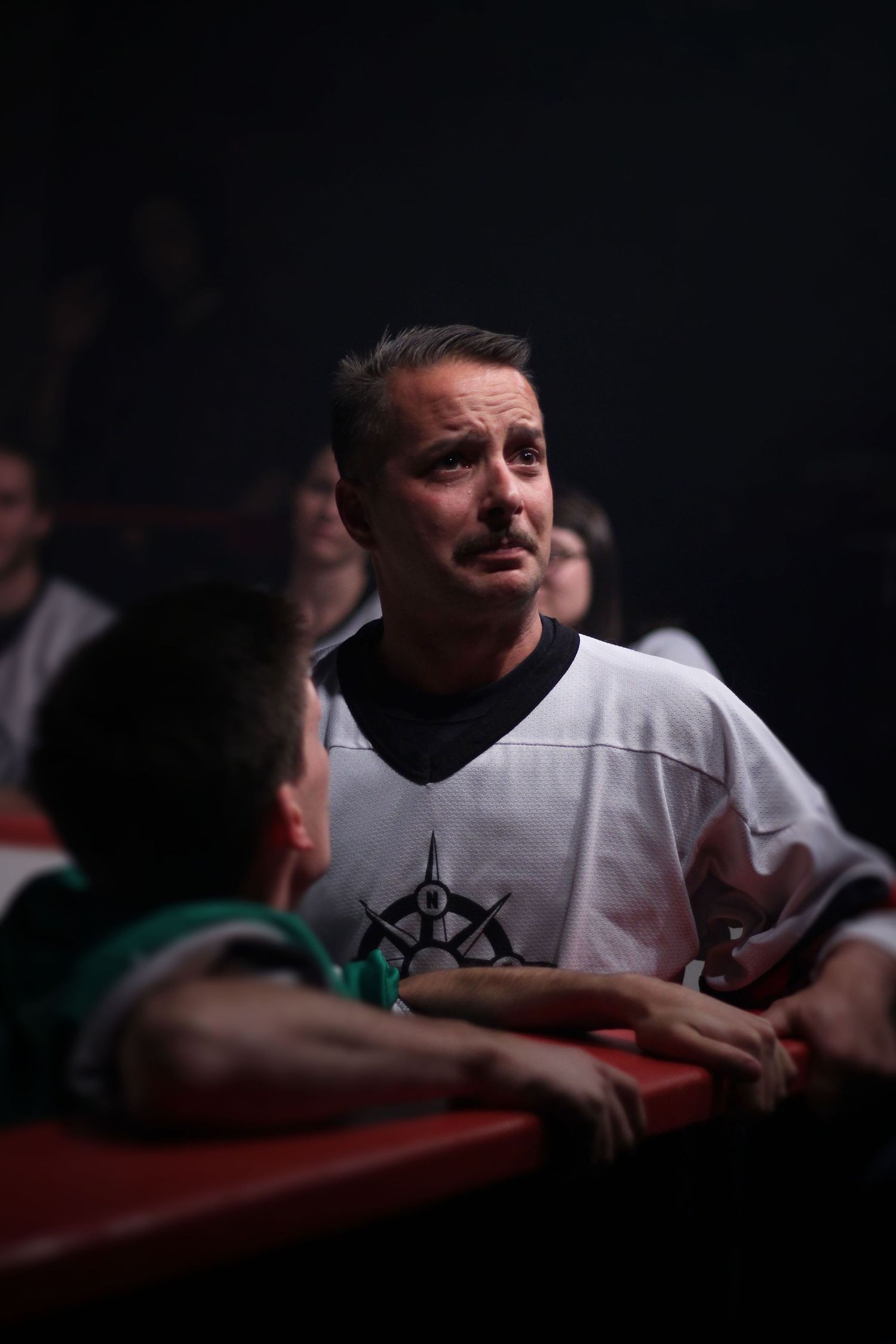 Man in white hockey jersey sitting in red theater seats looking upward with an expressive face.