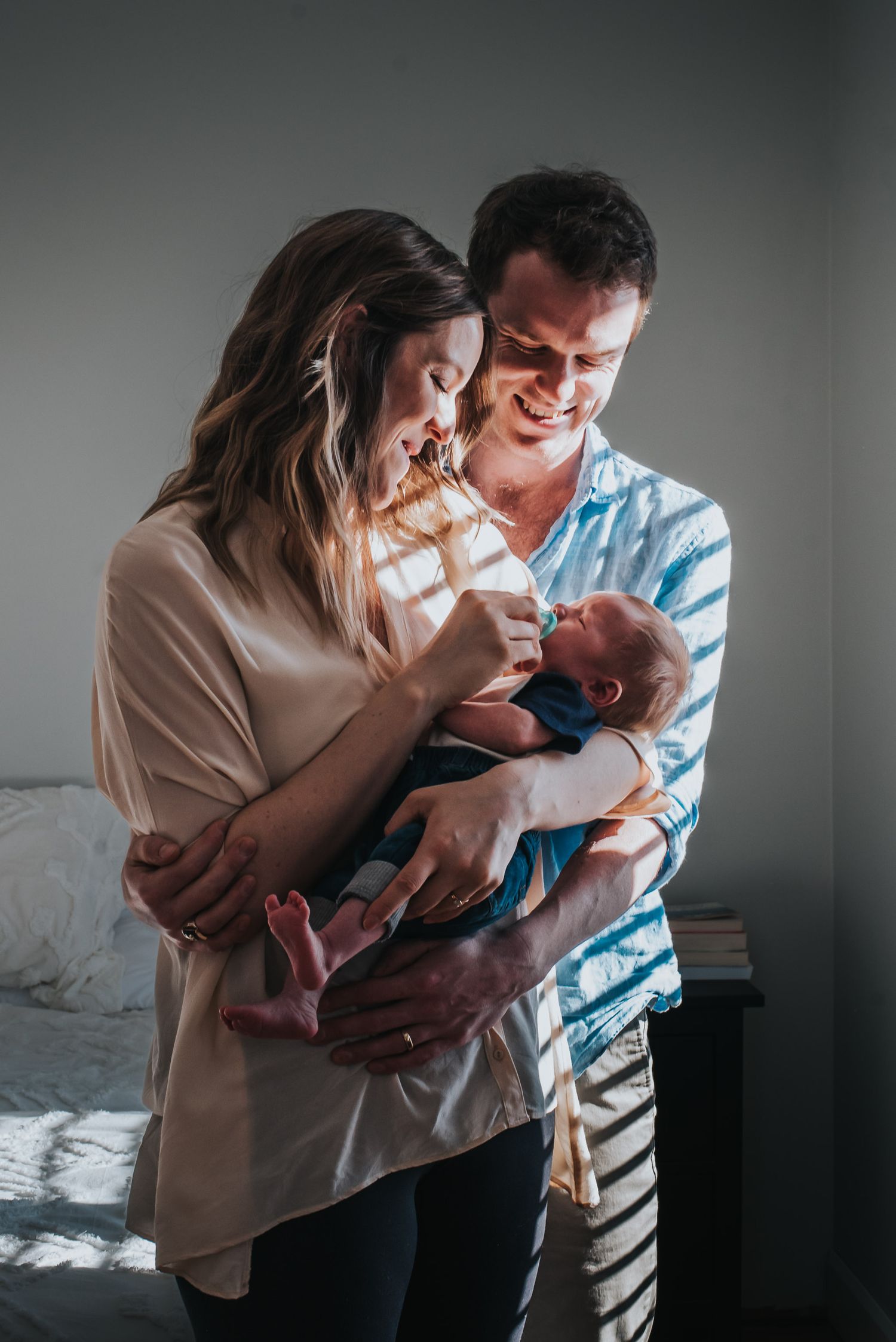 Window streams through slatted blinds onto a woman as she holds her newborn and is held by her husband.