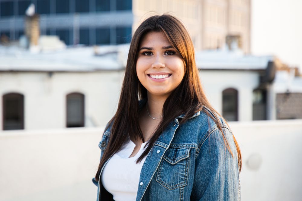 A person wearing a denim jacket and white top poses outdoors with urban architecture in the background.