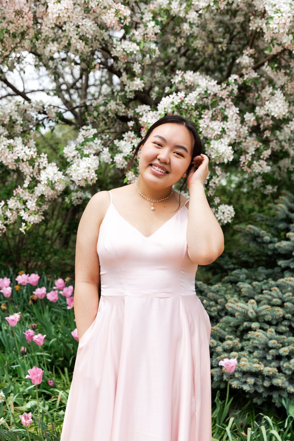 A person in a pink dress poses in front of blooming white flowers and pink tulips in a garden setting.