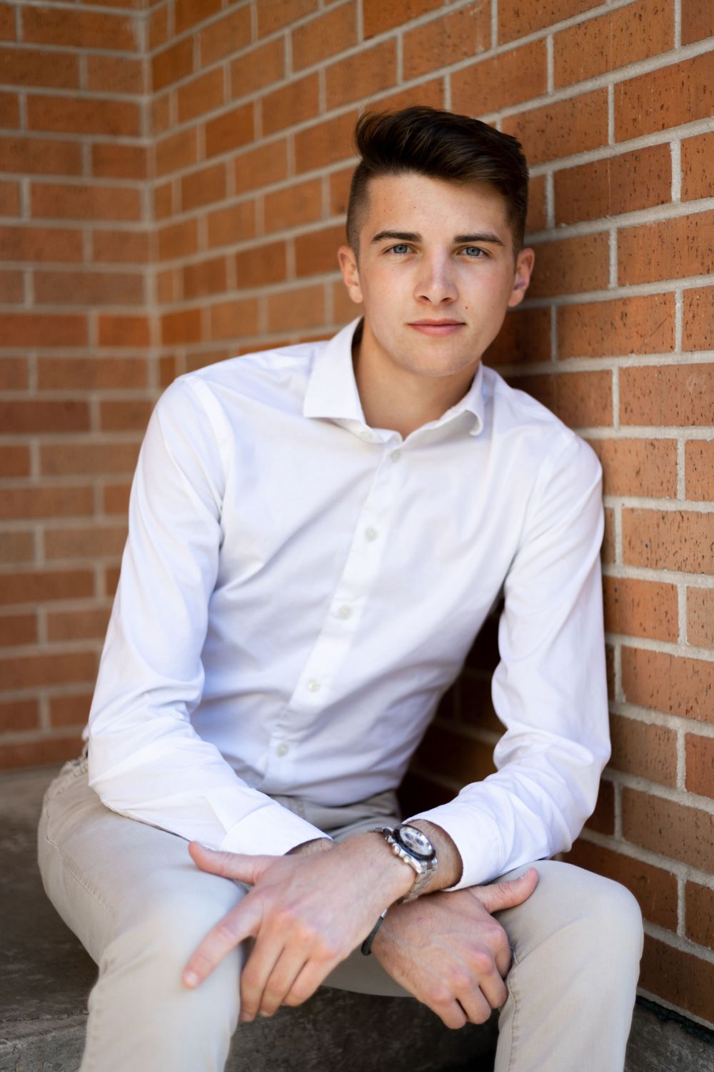 A person in a crisp white dress shirt and khaki pants sits casually against a brick wall.