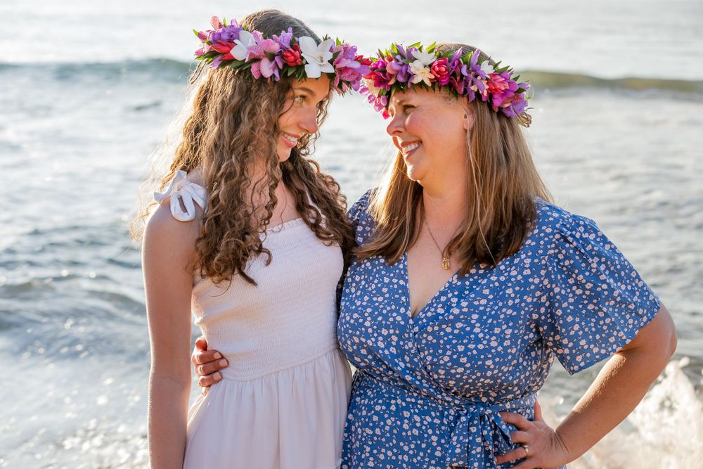 Two people wearing floral crowns share a joyful moment together on a beach at sunset.