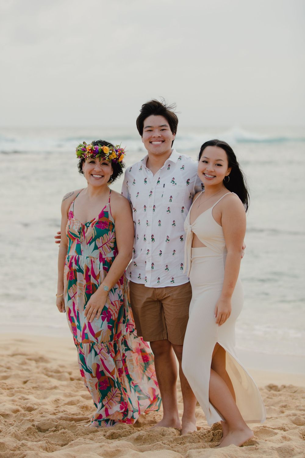 Three friends pose on a beach with one wearing a colorful flower crown and flowy dress.