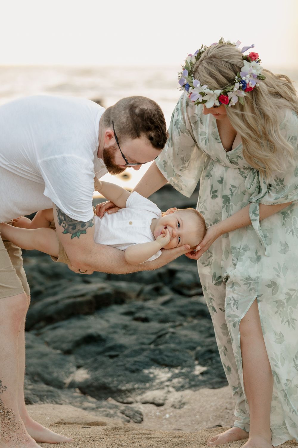 Parents playing with laughing baby on rocky beach during sunset photoshoot.
