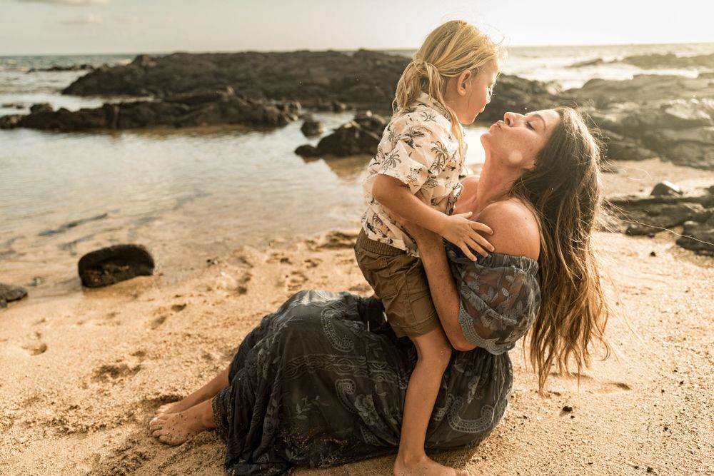 Intimate family moment captured on sandy beach with ocean rocks at sunset.