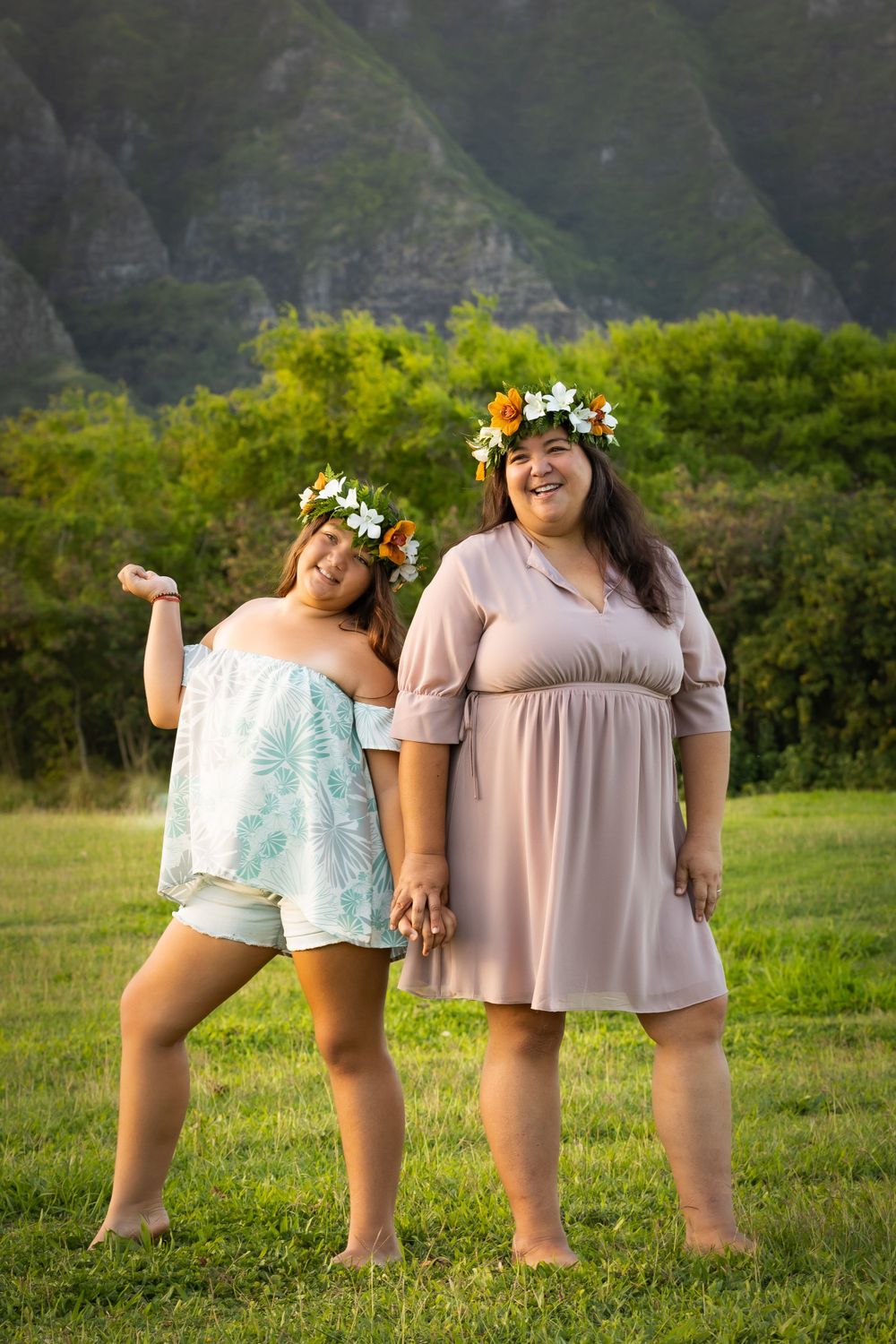 Two women wearing flower crowns pose joyfully in front of lush Hawaiian mountains.