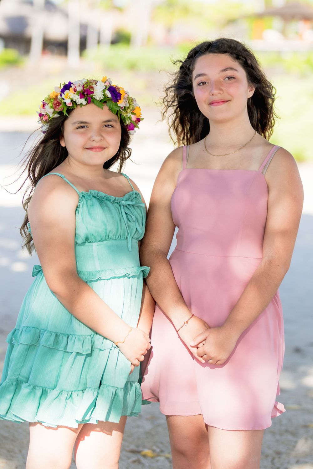 Two young people in summer dresses, one mint green with a floral crown and one pink, pose together outdoors.
