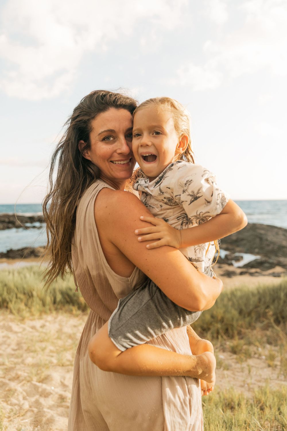 Parent and child share laughter while embracing on grassy coastal overlook.