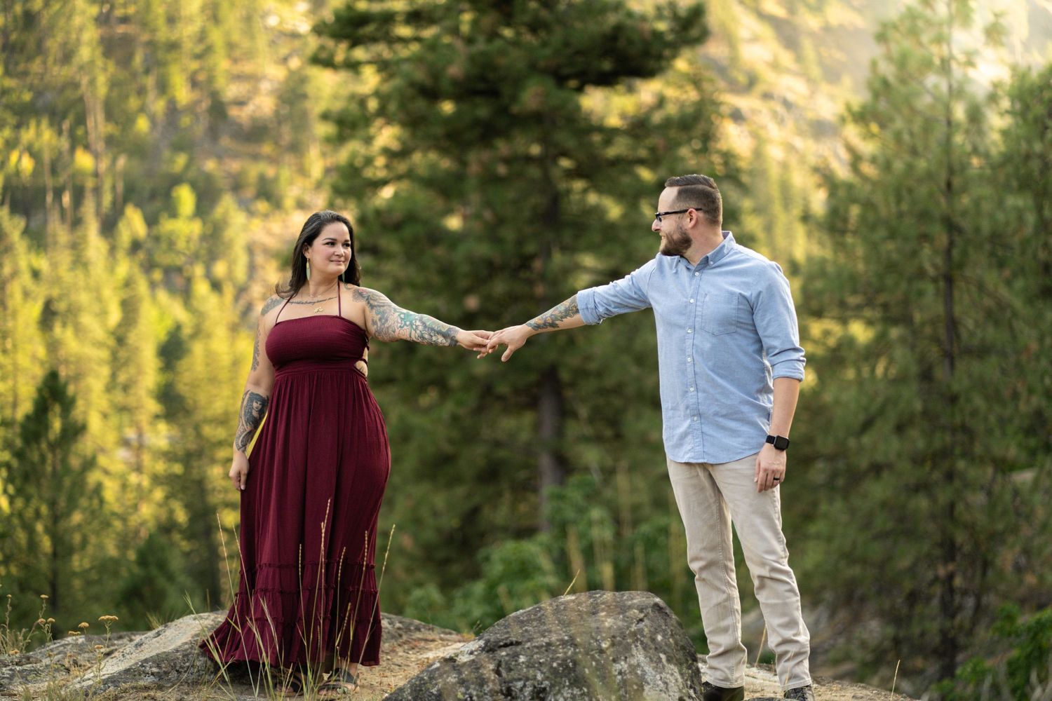 Couple holding hands while standing on rocky mountain overlook with forest and peaks in golden light.