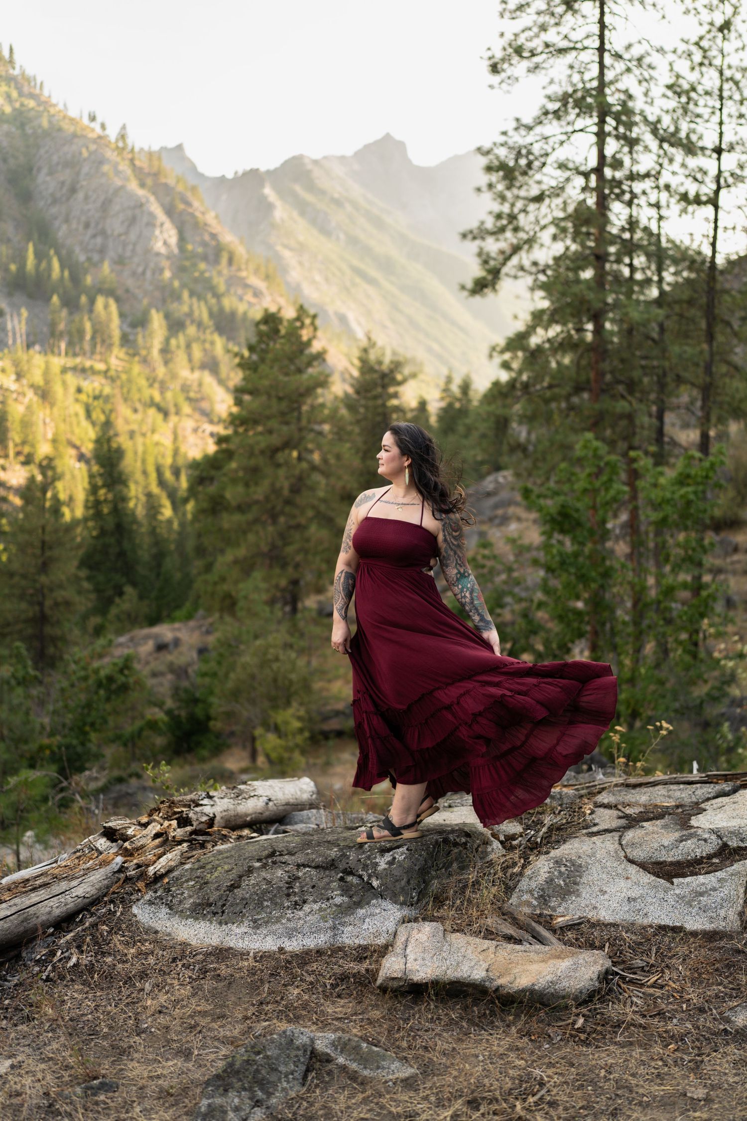 Woman in flowing burgundy dress stands on rocky overlook with dramatic mountain scenery behind.