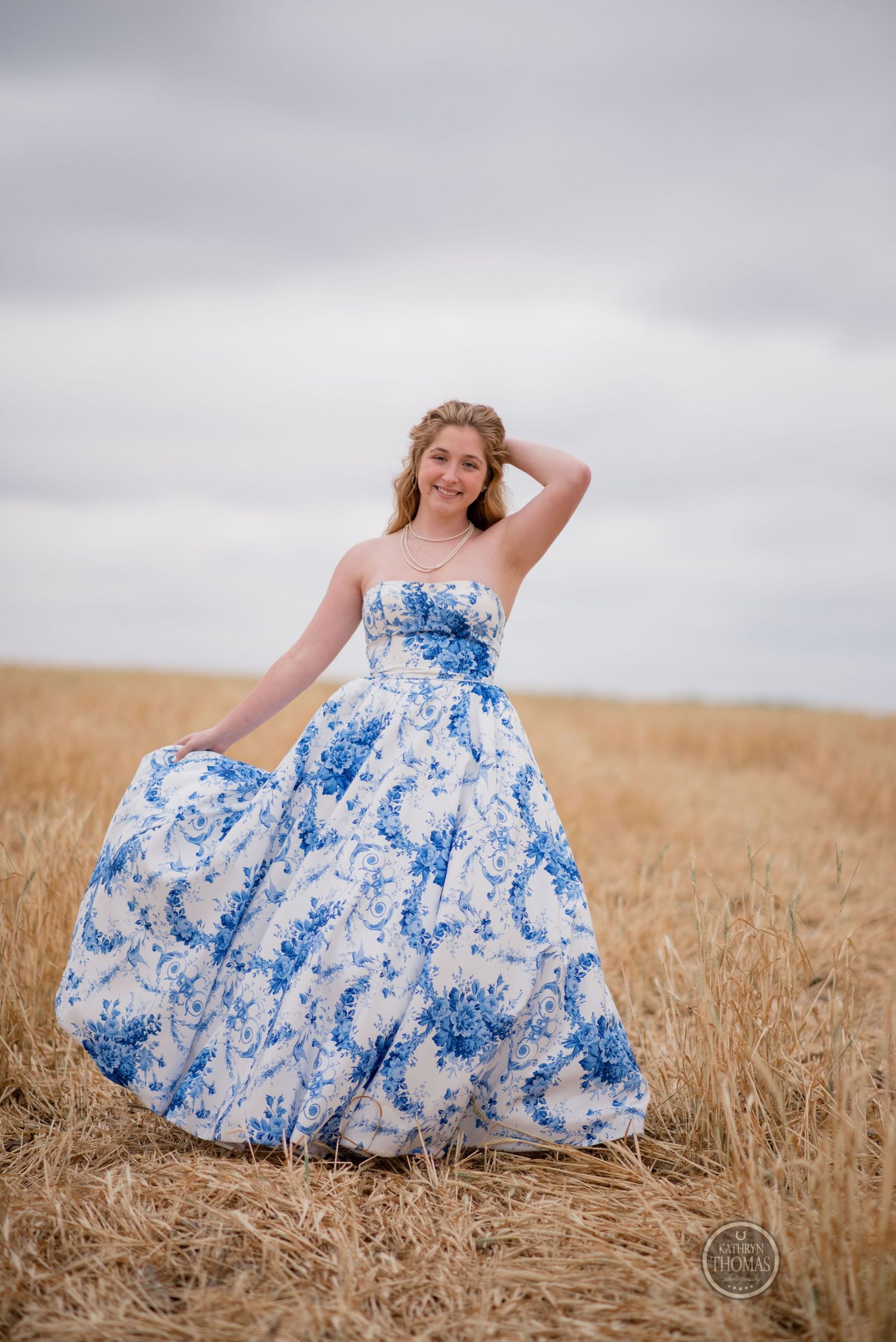 Carefree pose in a stunning blue and white ballgown against golden field and overcast sky.
