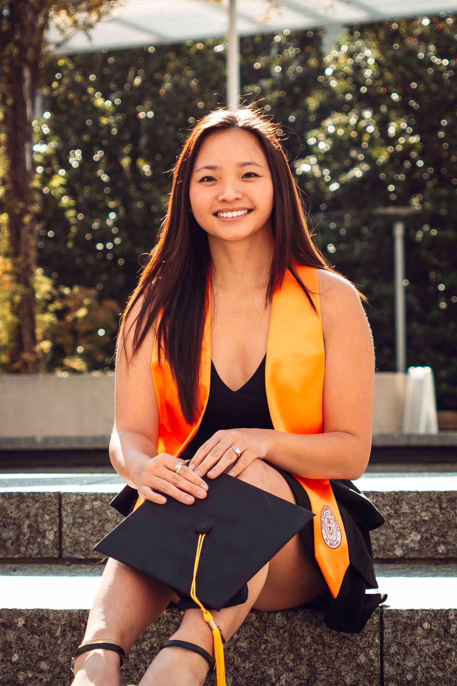 Graduate in orange stole sitting outdoors with diploma during graduation photoshoot.