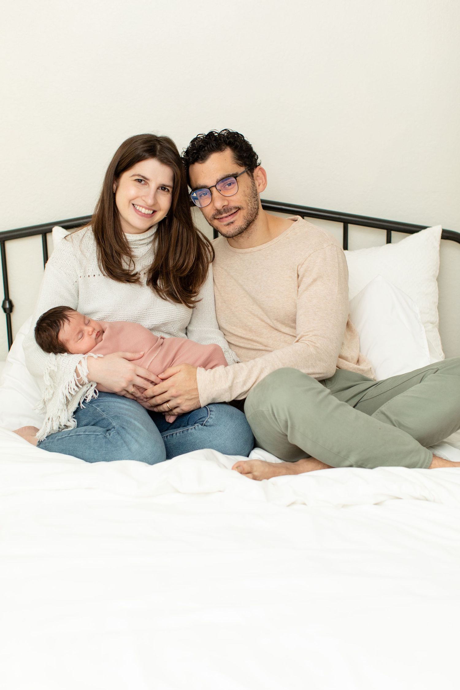 A couple relaxes on a bed with their newborn baby in a cozy bedroom setting.