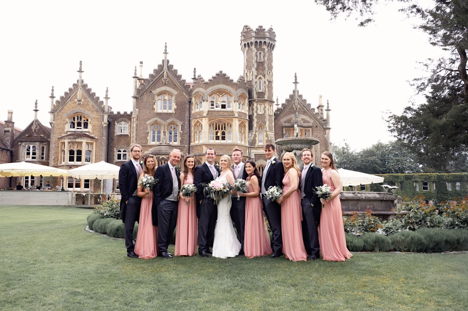 Wedding party poses in front of grand Gothic revival mansion with bridesmaids in pink dresses.