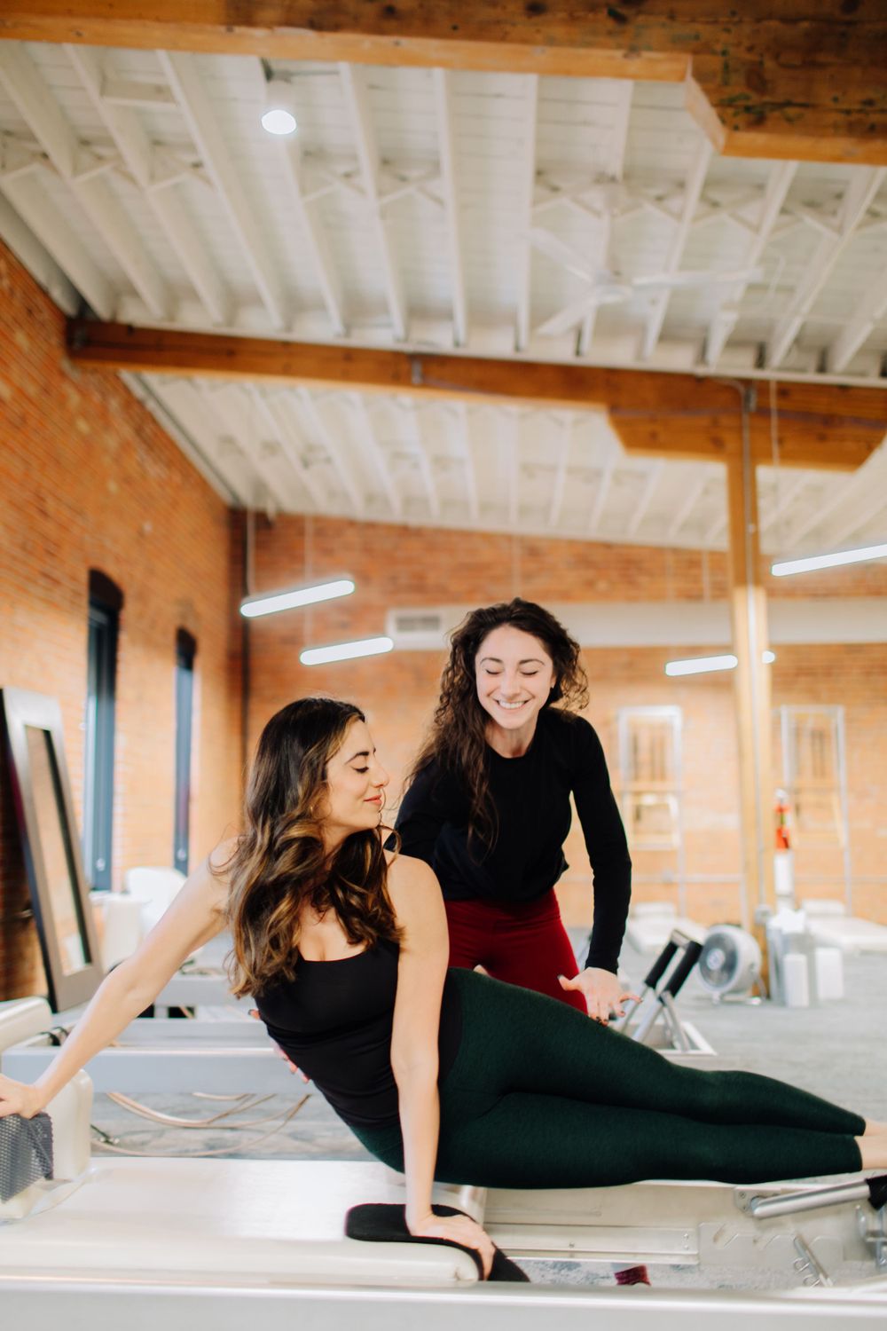 Two people practicing Pilates exercises on reformer machines in a brick-walled studio.