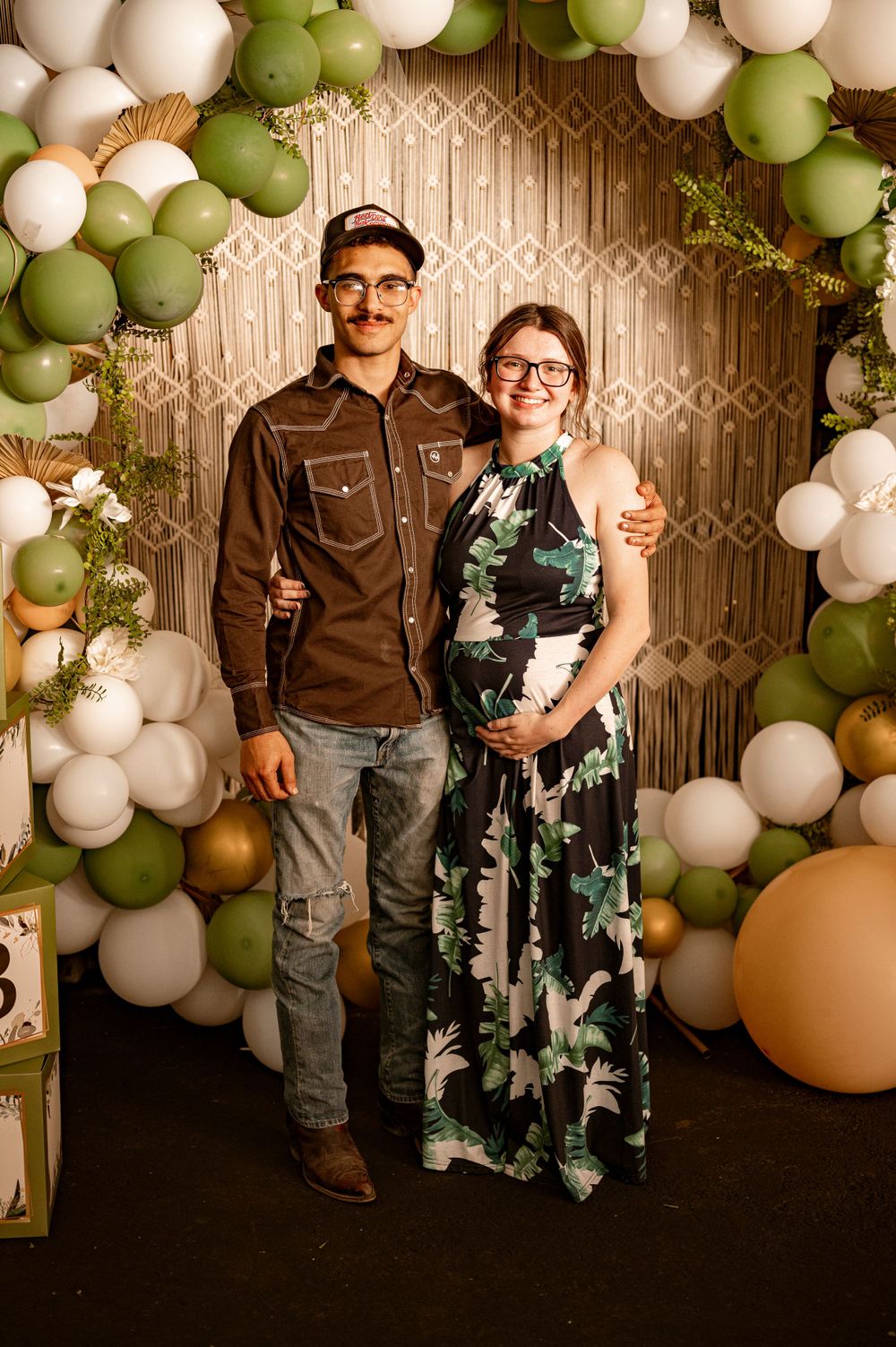 A couple poses under an elegant balloon arch with white and sage green balloons against a patterned backdrop.