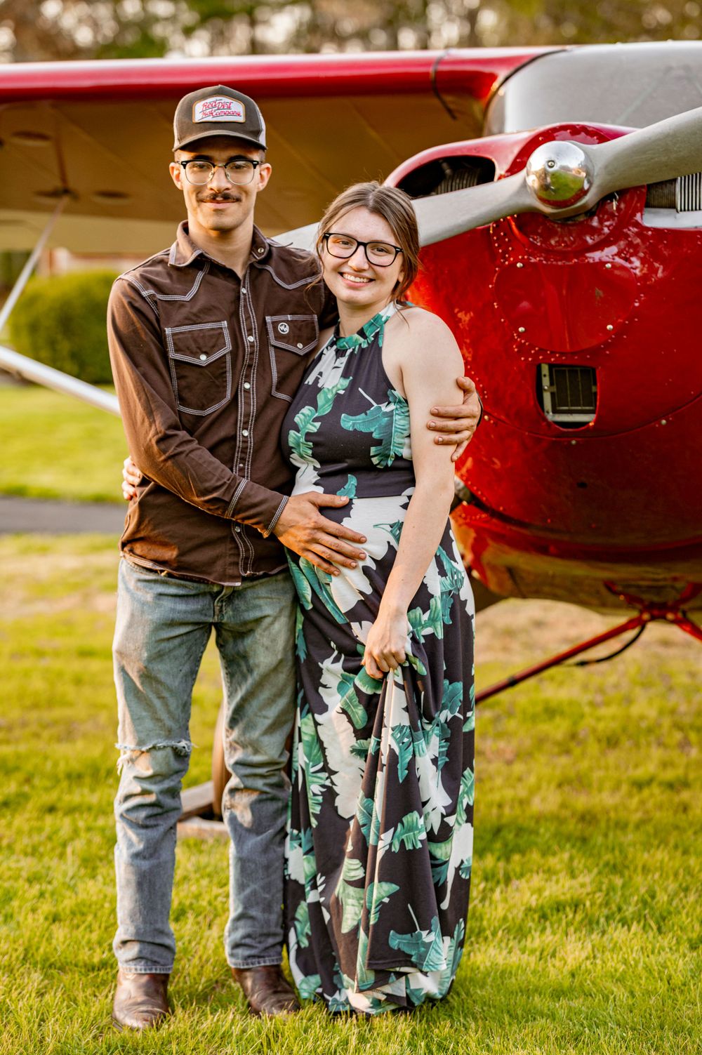 A couple stands in front of a red vintage airplane on a grassy field.