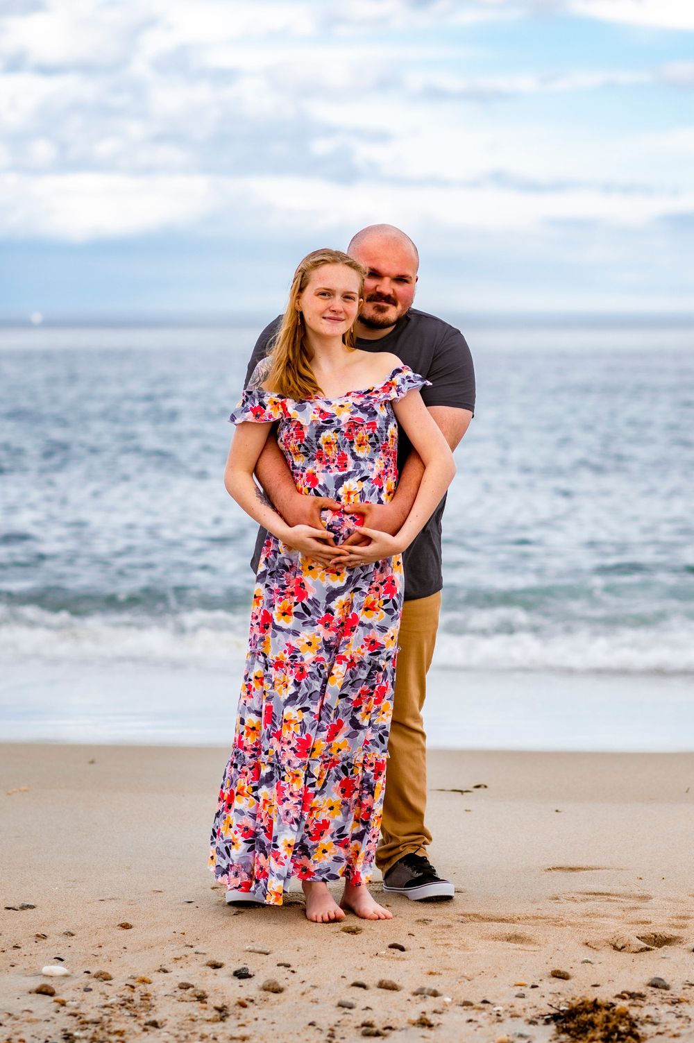 Couple poses on a beach with ocean waves and cloudy sky in the background.