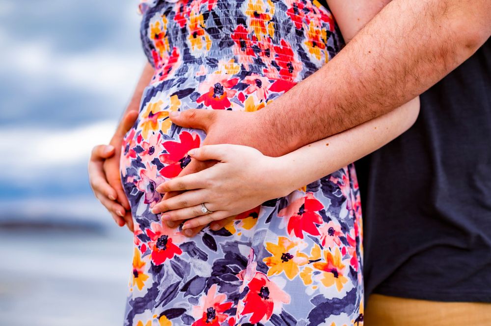Close-up of hands cradling a baby bump in a colorful floral dress on the beach.